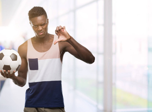 Молодий афроамериканець людиною проведення футбол футбольний м'яч з сердитися обличчя, ознака від'ємного числа показує неприязнь з великі пальці вниз, відмова концепції - Фото, зображення