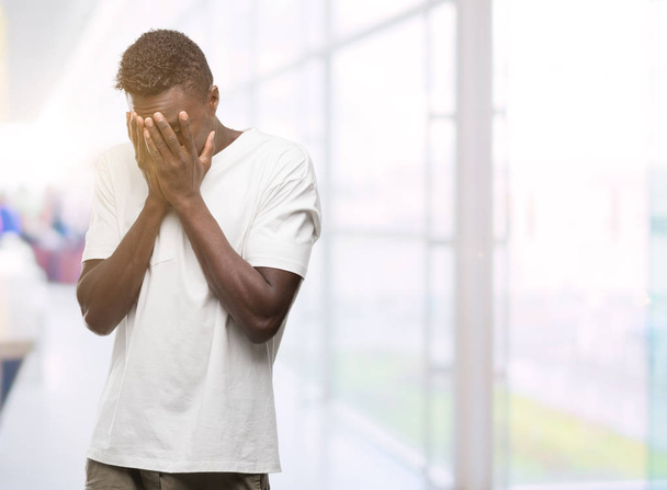 Joven hombre afroamericano vistiendo camiseta blanca con expresión triste cubriendo la cara con las manos mientras llora. Concepto de depresión
. - Foto, imagen