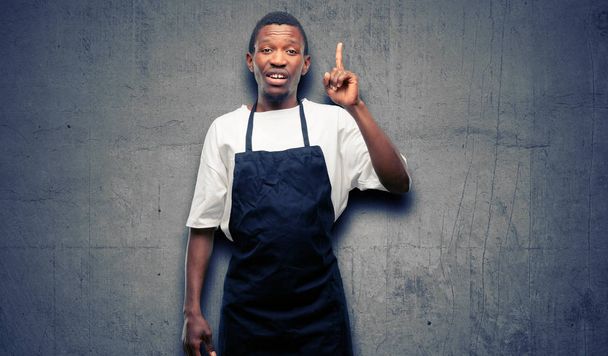 African man shop owner wearing apron happy and surprised cheering expressing wow gesture pointing up - Foto, afbeelding