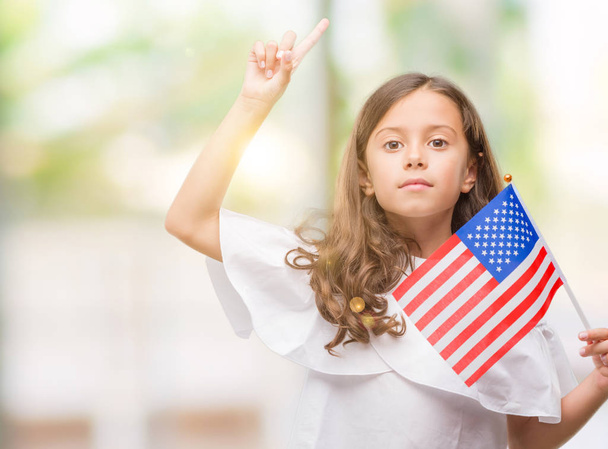 Menina hispânica morena segurando bandeira dos Estados Unidos da América surpreendida com uma ideia ou pergunta apontando o dedo com o rosto feliz, número um
 - Foto, Imagem