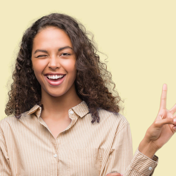 Jeune femme d'affaires hispanique souriant avec le visage heureux clin d'oeil à la caméra faisant signe de victoire. Numéro deux
. - Photo, image