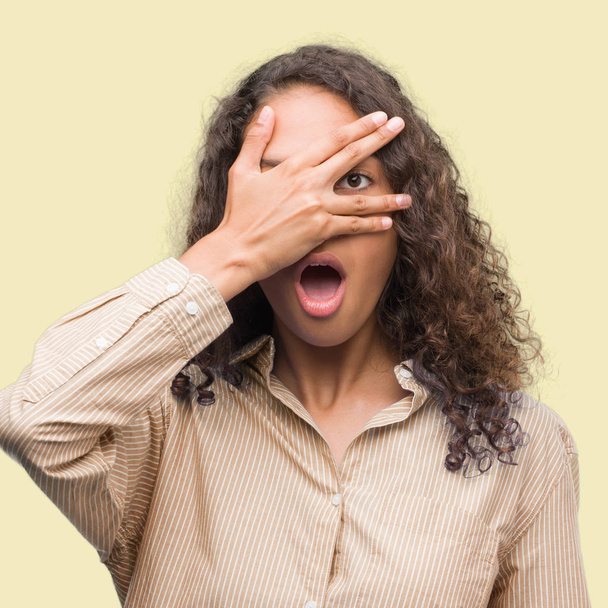 Young hispanic business woman peeking in shock covering face and eyes with hand, looking through fingers with embarrassed expression. - Photo, Image