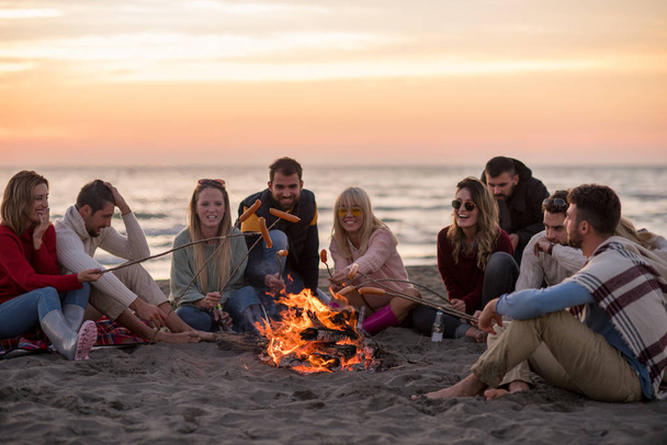 Group of young friends sitting by the fire at autumn beach, grilling sausages and drinking beer, talking and having fun - Фото, зображення