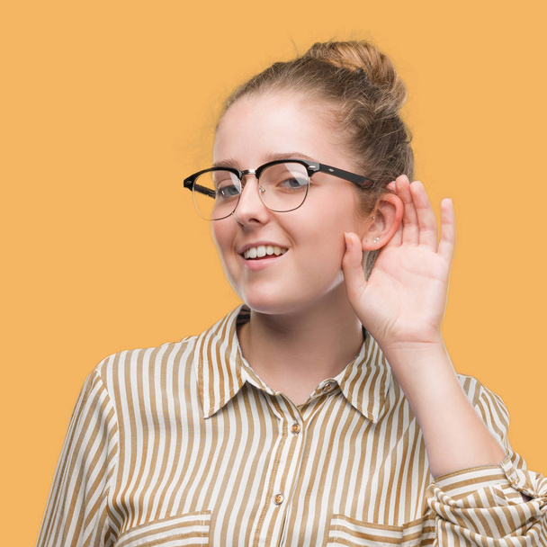 Jonge blonde zakenvrouw glimlachend met hand over oor luisteren een hoorzitting gerucht of roddel. Doofheid concept. - Foto, afbeelding