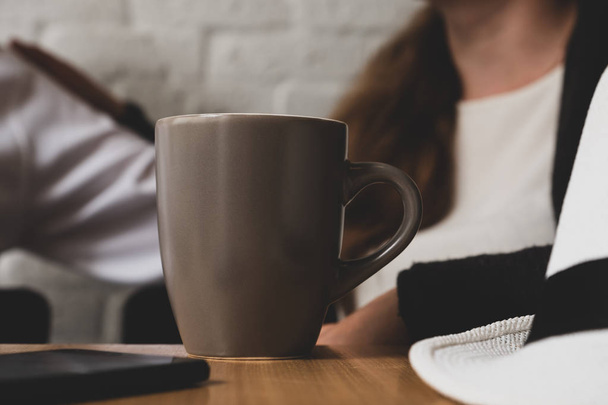 Auf einem Tisch steht eine Tasse Kaffee. Im Hintergrund ist der Mann und die Frau sichtbar. vor einem Becher auf einem Tischtelefon und einem Hut liegt. das Bild ist getönt - Foto, Bild