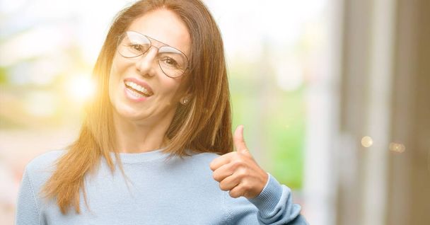 Middelbare leeftijd vrouw dragen van wollen trui en koele glazen glimlachend in grote lijnen weergegeven: duimschroef opwaarts gebaar camera, expressie van en goedkeuring - Foto, afbeelding
