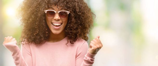 Αφρικανική αμερικανική γυναίκα που φοράει ροζ γυαλιά ηλίου, γιορτάζοντας έκπληκτος και κατάπληκτος για την επιτυχία με τα όπλα υπερυψωμένο και ανοικτά μάτια. Νικητής έννοια. - Φωτογραφία, εικόνα