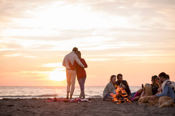 Fiatal pár élvezi a barátaival körül tábortűz a strandon Naplementekor sört iszik - Fotó, kép