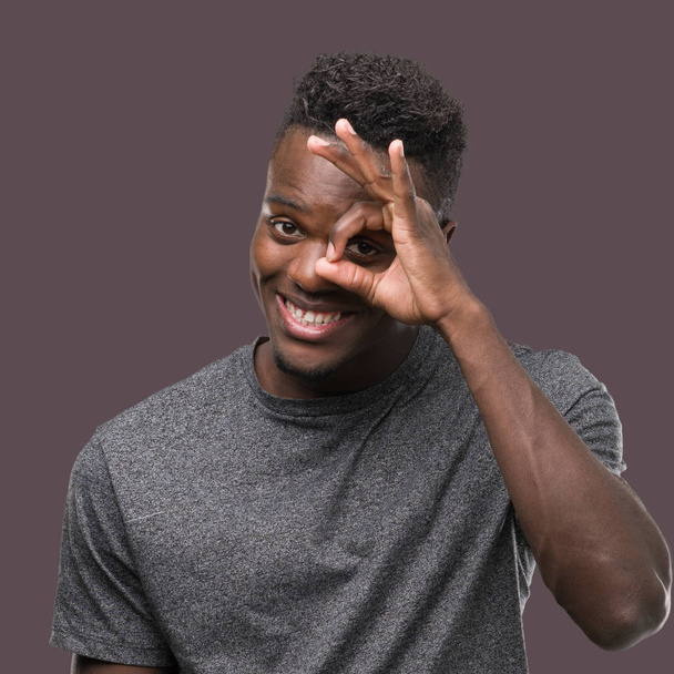 Молодой африканский американец в серой футболке делает жест с улыбкой на руке, глаза смотрят сквозь пальцы со счастливым лицом
. - Фото, изображение