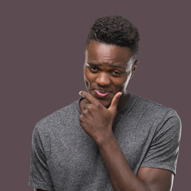 Νεαρός αφρικανική αμερικανική άνθρωπος που φορώντας γκρι t-shirt που αναζητούν αυτοπεποίθηση στη φωτογραφική μηχανή με χαμόγελο με σταυρωμένα χέρια και χέρι σηκωμένο στο πηγούνι. Σκέφτεστε θετικά. - Φωτογραφία, εικόνα