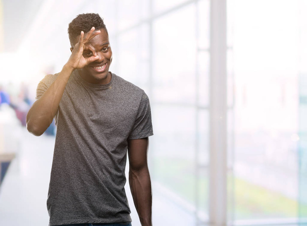 Jonge african american man die grijs t-shirt doen ok gebaar met hand glimlachen, oog kijken door vingers met blij gezicht. - Foto, afbeelding