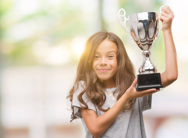 Brunette hispanic girl holding a trophy with a happy face standing and smiling with a confident smile showing teeth - Valokuva, kuva