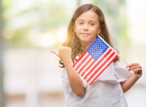 Menina hispânica morena segurando bandeira dos Estados Unidos da América apontando e mostrando com o polegar para o lado com o rosto feliz sorrindo
 - Foto, Imagem