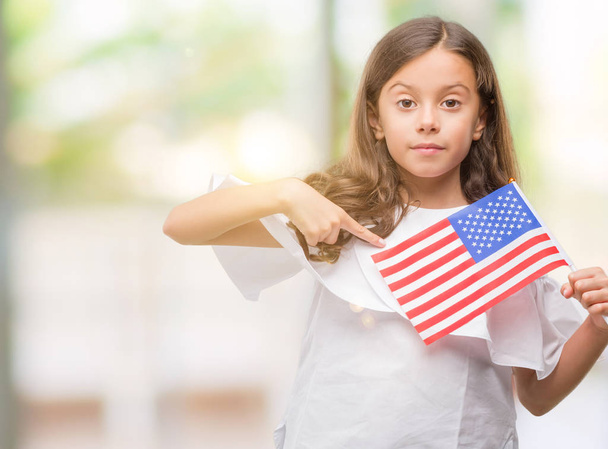 Barna hispán lány gazdaság meglepetés arc mutatóujjal Egyesült Államok zászlaja, hogy maga - Fotó, kép