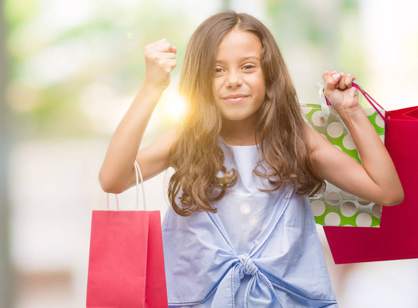 Brunette hispanic girl holding shopping bags screaming proud and celebrating victory and success very excited, cheering emotion - Foto, afbeelding