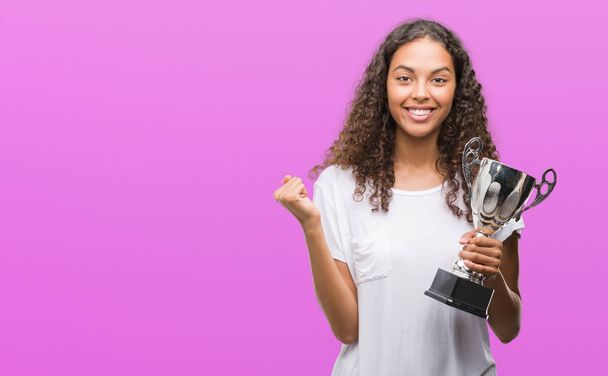 Young hispanic woman holding trophy screaming proud and celebrating victory and success very excited, cheering emotion - Photo, Image