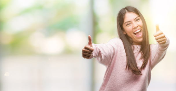 Young beautiful hispanic woman wearing a sweater approving doing positive gesture with hand, thumbs up smiling and happy for success. Looking at the camera, winner gesture. - Photo, Image
