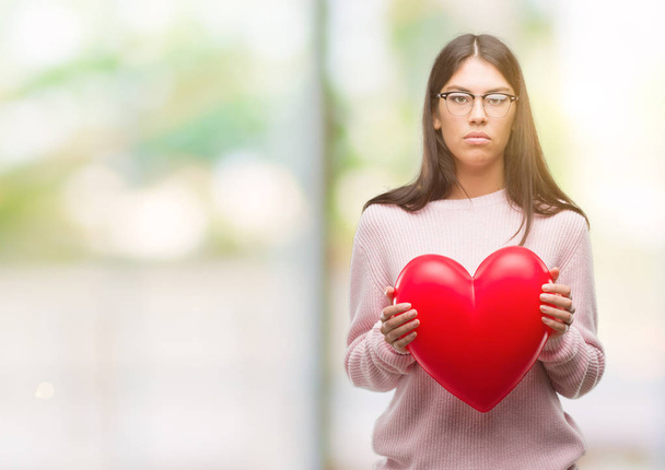 Young hispanic woman holding heart in love with a confident expression on smart face thinking serious - Fotoğraf, Görsel