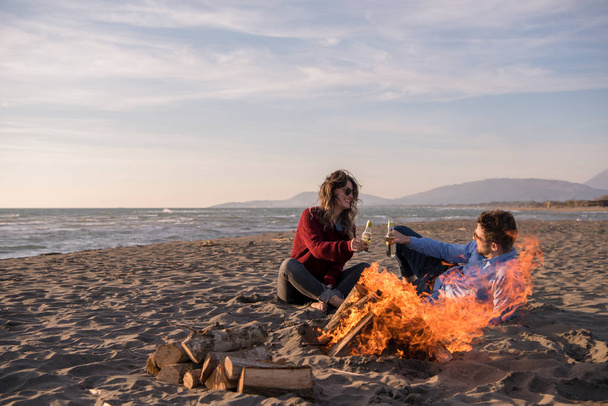 Young Couple Relaxing By The Fire, Drinking A Beer Or A Drink From The Bottle on the beach at autumn day - Foto, immagini