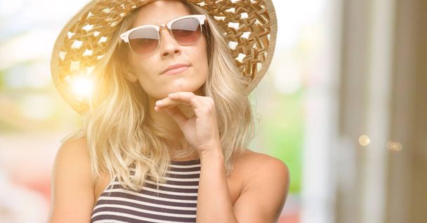 Young woman using sunglasses wearing summer hat thinking and looking up expressing doubt and wonder - Photo, Image