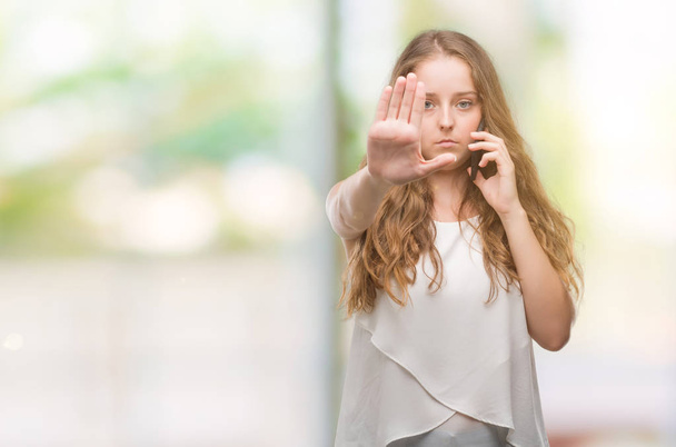 Young blonde woman using smartphone with open hand doing stop sign with serious and confident expression, defense gesture - Photo, Image