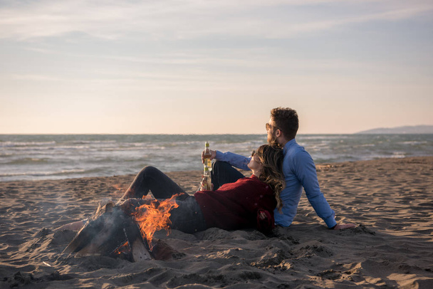 Young Couple Relaxing By The Fire, Drinking A Beer Or A Drink From The Bottle on the beach at autumn day - Foto, Imagem