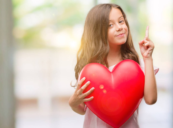Брюнетка латиноамериканського дівчина Холдинг червоне серце здивований ідеї або питання вказівним пальцем з щасливим обличчям, номер один - Фото, зображення