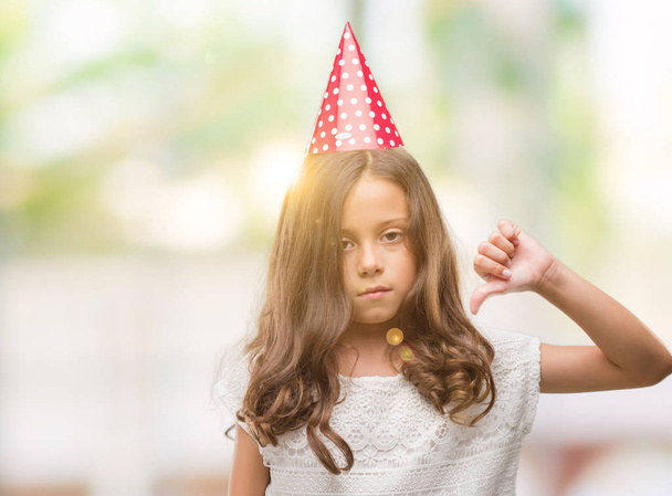 Μελαχρινή κορίτσι Ισπανόφωνος φοράει καπέλο γενεθλίων με θυμωμένο πρόσωπο, αρνητικό πρόσημο δείχνει απέχθεια με αντίχειρες προς τα κάτω, απόρριψη έννοια - Φωτογραφία, εικόνα