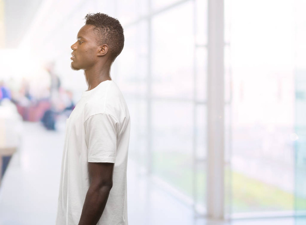 junger afrikanisch-amerikanischer Mann trägt weißes T-Shirt, das zur Seite schaut, entspannte Profilpose mit natürlichem Gesicht und selbstbewusstem Lächeln. - Foto, Bild