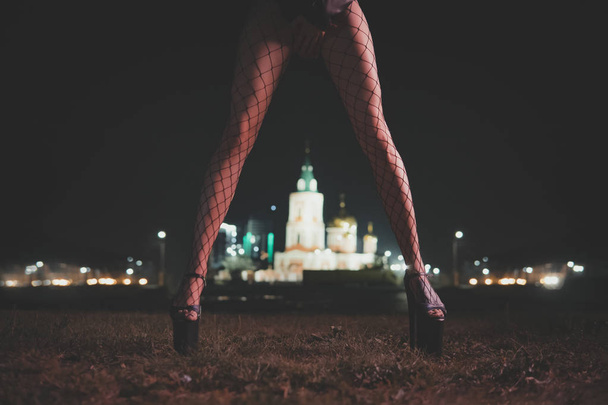 girl shyly covers her hands with an intimate part of the body. Slender female legs stand against the night city on the street. - Photo, Image