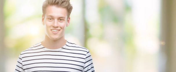 Jovem homem loiro bonito vestindo listras marinheiro t-shirt com um rosto feliz de pé e sorrindo com um sorriso confiante mostrando dentes
 - Foto, Imagem