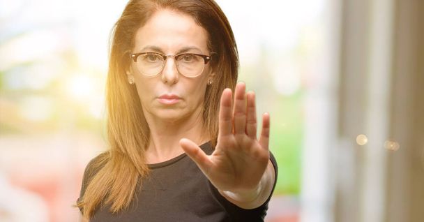 Mujer de mediana edad vistiendo camisa negra y gafas molestas con mala actitud haciendo señal de stop con la mano, diciendo no, expresando seguridad, defensa o restricción, tal vez empujando
 - Foto, imagen