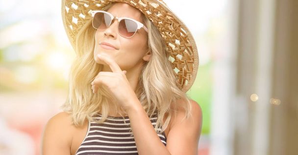 Mujer joven usando gafas de sol con sombrero de verano pensando y mirando hacia arriba expresando dudas y maravillas
 - Foto, imagen