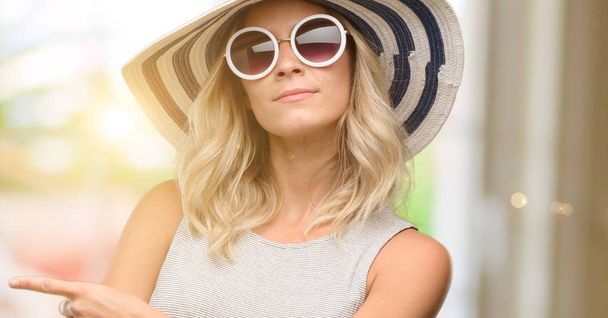 Νεαρή γυναίκα που φοράει γυαλιά ηλίου και το καλοκαιρινό καπέλο δείχνει μακριά πλευρά με το δάχτυλο - Φωτογραφία, εικόνα