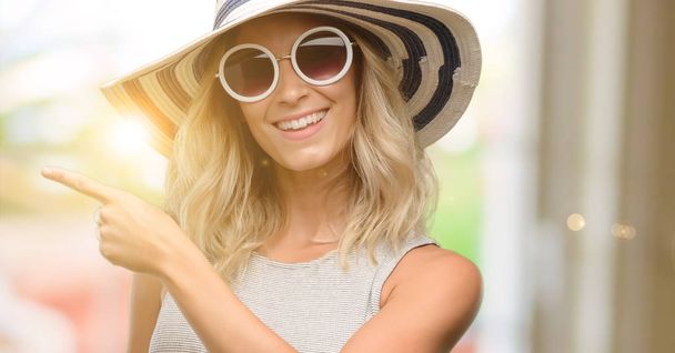 Young woman wearing sunglasses and summer hat pointing away side with finger - Photo, Image