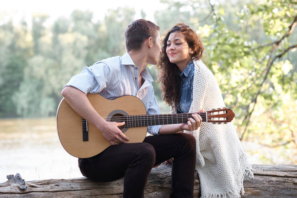 young couple sitting on a log by the river and playing guitar, summer nature, bright sunlight, shadows and green leaves, romantic feelings - Foto, Imagen