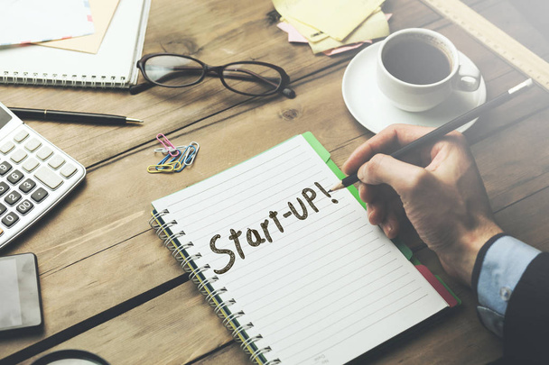man has written start-up text on notebook  with stationary and coffee on wooden  table - Foto, Imagen