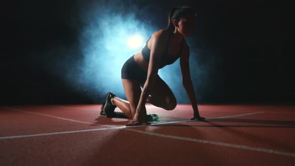 Atleta femenina sobre un fondo oscuro se prepara para correr el sprint a campo traviesa desde las almohadillas de la cinta sobre un fondo oscuro
. - Metraje, vídeo
