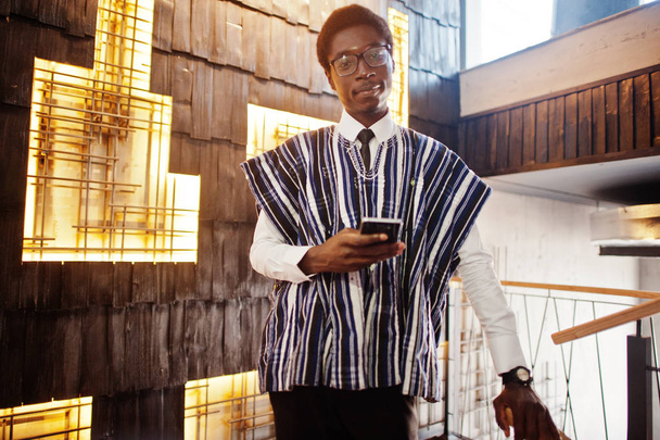 Afrikanischer Geschäftsmann in traditioneller Kleidung und Brille mit Handy in der Hand posiert in modernem Gebäude. - Foto, Bild