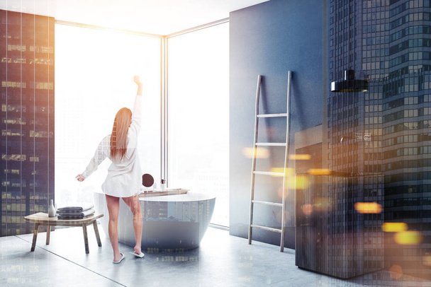 Woman in pajamas in gray wall bathroom corner with concrete floor, loft window, ladder, wood sink and bathtub. Spa, hotel luxury real estate. 3d rendering mock up toned image double exposure city - Foto, immagini