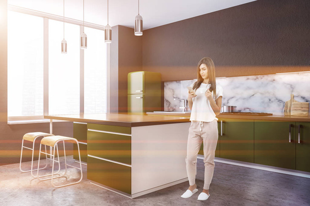 Woman with smartphone in modern marble wall kitchen corner with large windows, a concrete floor and green countertops with built in appliances. Island in the center 3d rendering mock up toned image - Foto, afbeelding