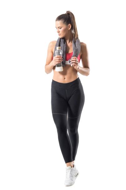Confident healthy sportswoman in sportswear with towel and glass water bottle looking down. Full body length portrait isolated on white background. - Foto, Bild