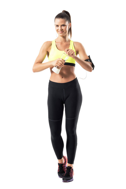 Sportif kadın sarı tişört ve su şişe açılış ve kameraya gülümseyen eğri uygun. Beyaz arka plan üzerinde izole tam vücut uzunluğu portre. - Fotoğraf, Görsel