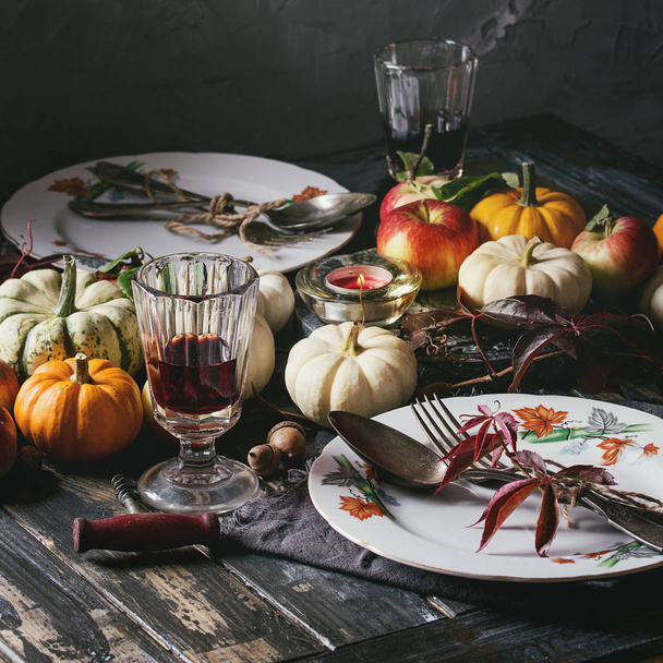 Autumn holiday table decoration setting with decorative pumpkins, apples, red leaves, empty plate with vintage cutlery, red wine, candle over wooden table. Rustic style. Square image - Photo, Image