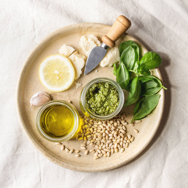 Traditional Basil pesto sauce in glass jar with ingredients above basil, olive oil, parmesan cheese, garlic, pine nuts, on ceramic plate over white linen cloth background. Flat lay, space. Square image - Foto, Imagem