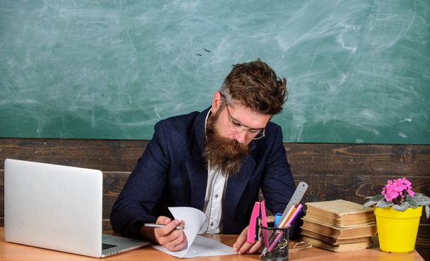 Check homework. Teacher bearded hipster with eyeglasses sit in classroom chalkboard background. Teacher sit desk with laptop. School teacher checking homework or test. Paperwork part of teachers life - Photo, Image