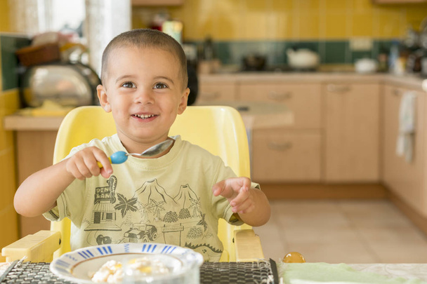 szczęśliwy chłopczyk jeść owsiankę. Mały chłopiec jedzenie śniadania na stole - Zdjęcie, obraz