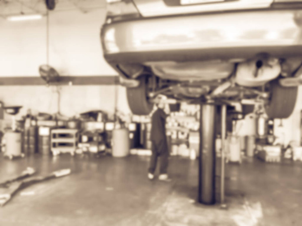 Vintage tone blurred mechanic changing oil below lifted car at auto shop in Texas, USA. Defocused background interior of modern oil change service station. Working technician at garage concept - Photo, Image