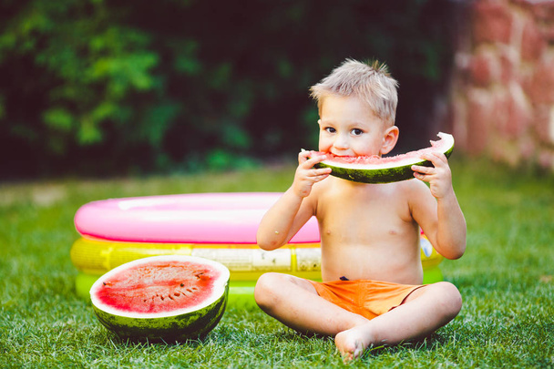 Theme summer seasonal food juicy watermelon berry. small funny Caucasian boy child two years sitting backyard green grass, lawn and eating piece watermelon holding hands. Naked in shorts and barefoot. - Photo, Image