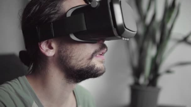 Young bearded hipster man using his VR headset display with headphones for virtual reality game or watching the 360 video while sitting on sofa at home in the living room. VR Technology. - Πλάνα, βίντεο